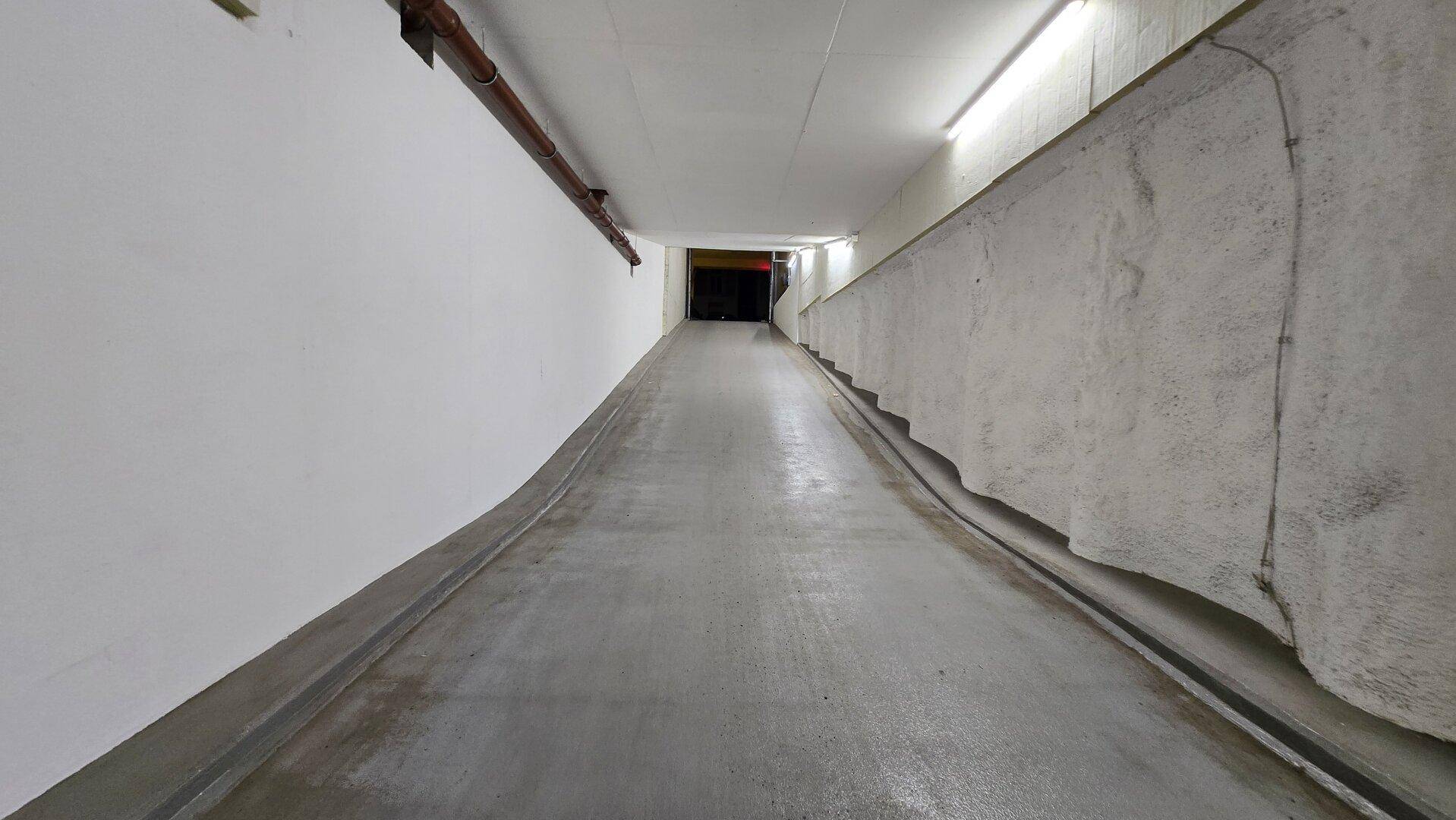 Lange, abfallende Betonrampe führt in eine Tiefgarage mit weißen Wänden und sichtbaren Versorgungsleitungen.