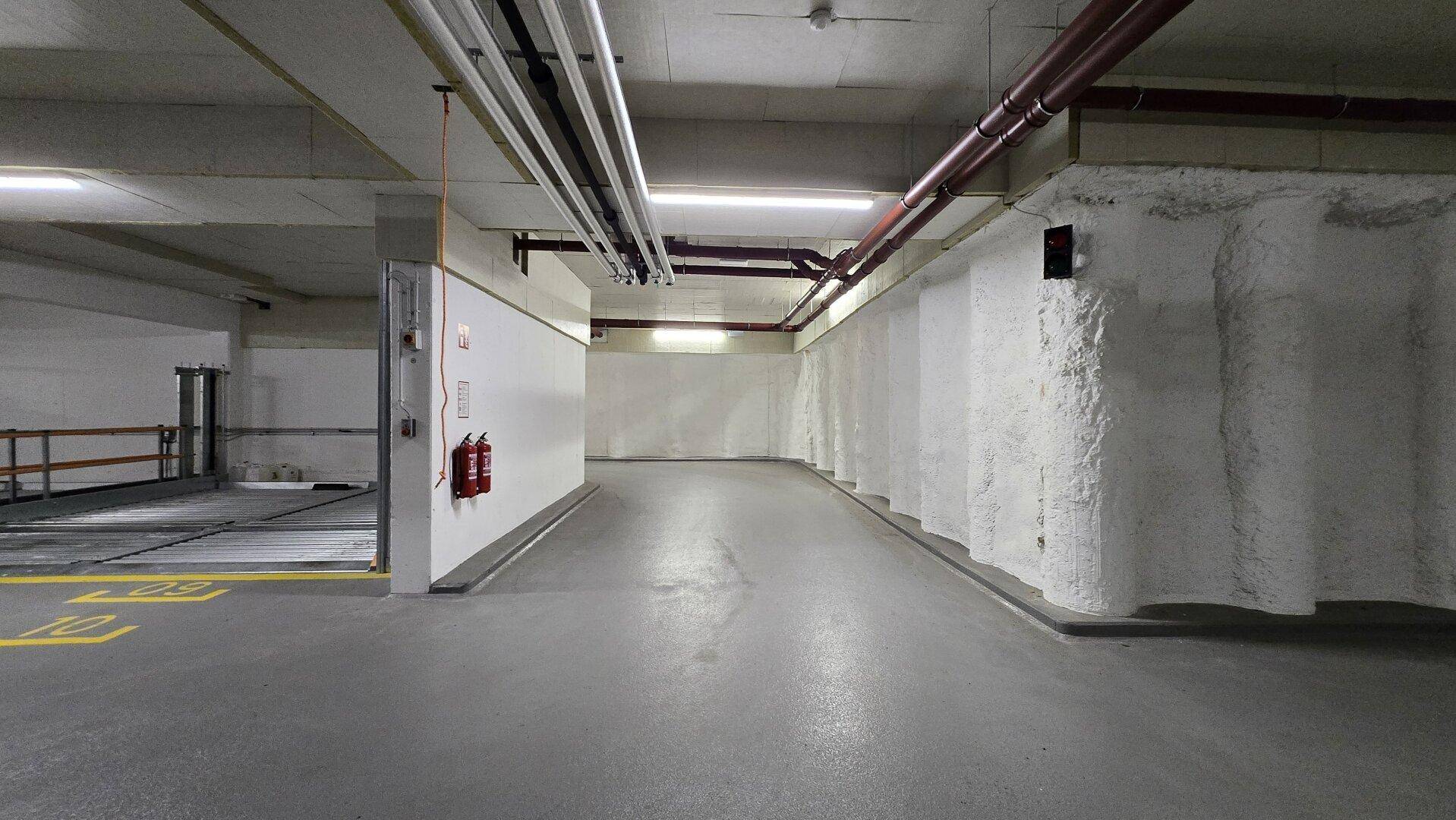 Geräumige Tiefgarage mit Betonboden, weißen Wänden, roten Rohren an der Decke und einem Feuerlöscher.