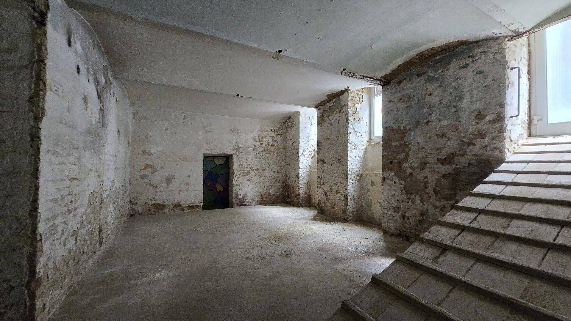 Kellerraum mit unverputzten Ziegelwänden, Betonboden und einer Holzrampe, die nach oben führt.