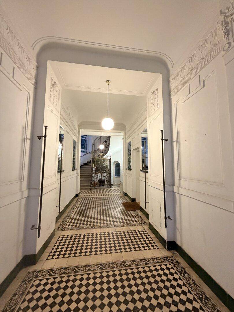 Historischer Flur mit Schachbrettmuster-Fliesenboden, hohen Decken und einer eleganten Treppe im Hintergrund.