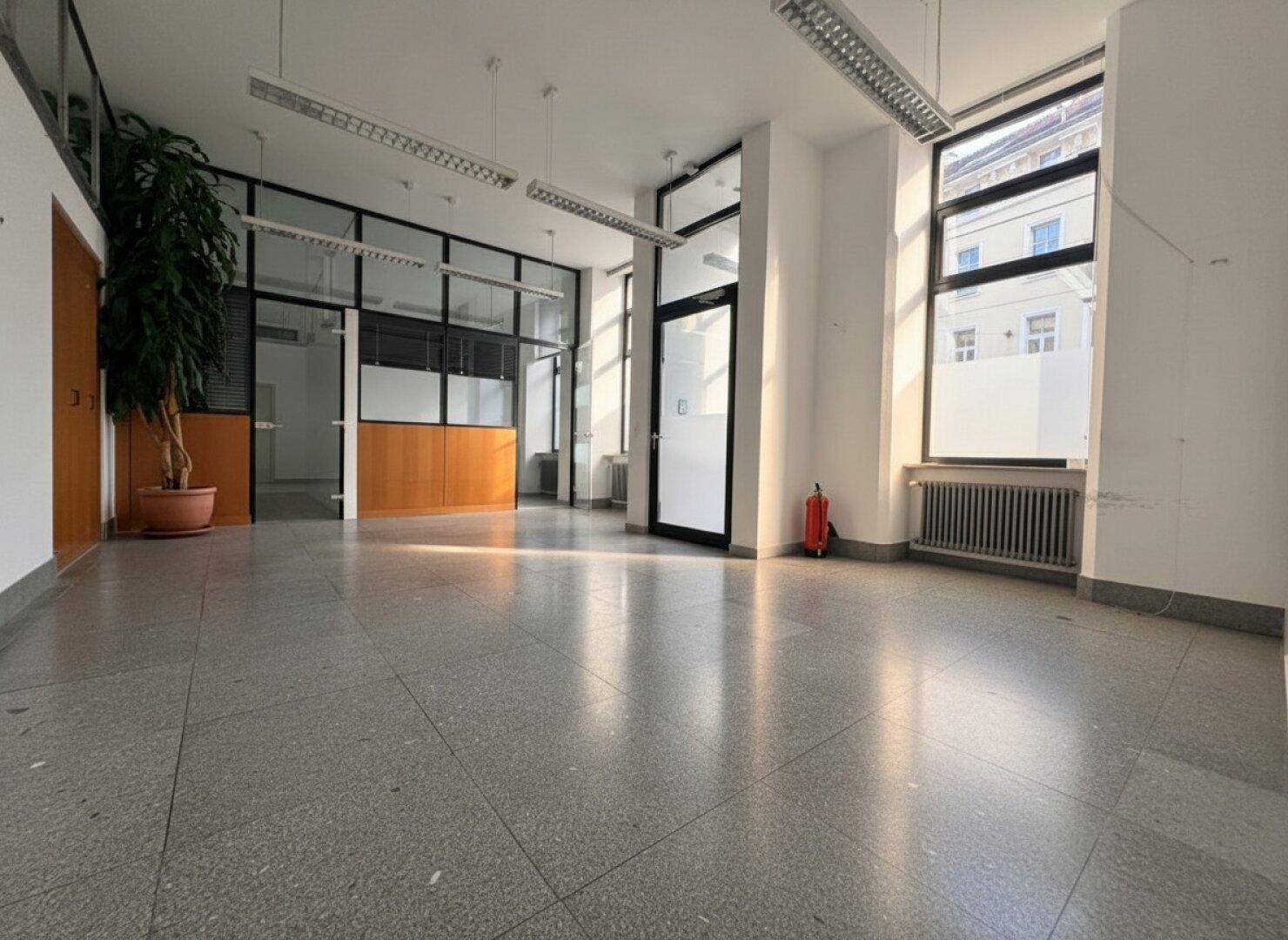 Geräumiger Bürobereich mit Terrazzoboden, Glaswänden und einem großen Fenster, das viel Licht hereinlässt.