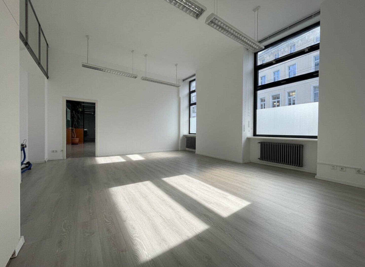 Leerer Büroraum mit hellem Laminatboden, hohen Decken und großen Fenstern, die viel Sonnenlicht hereinlassen.