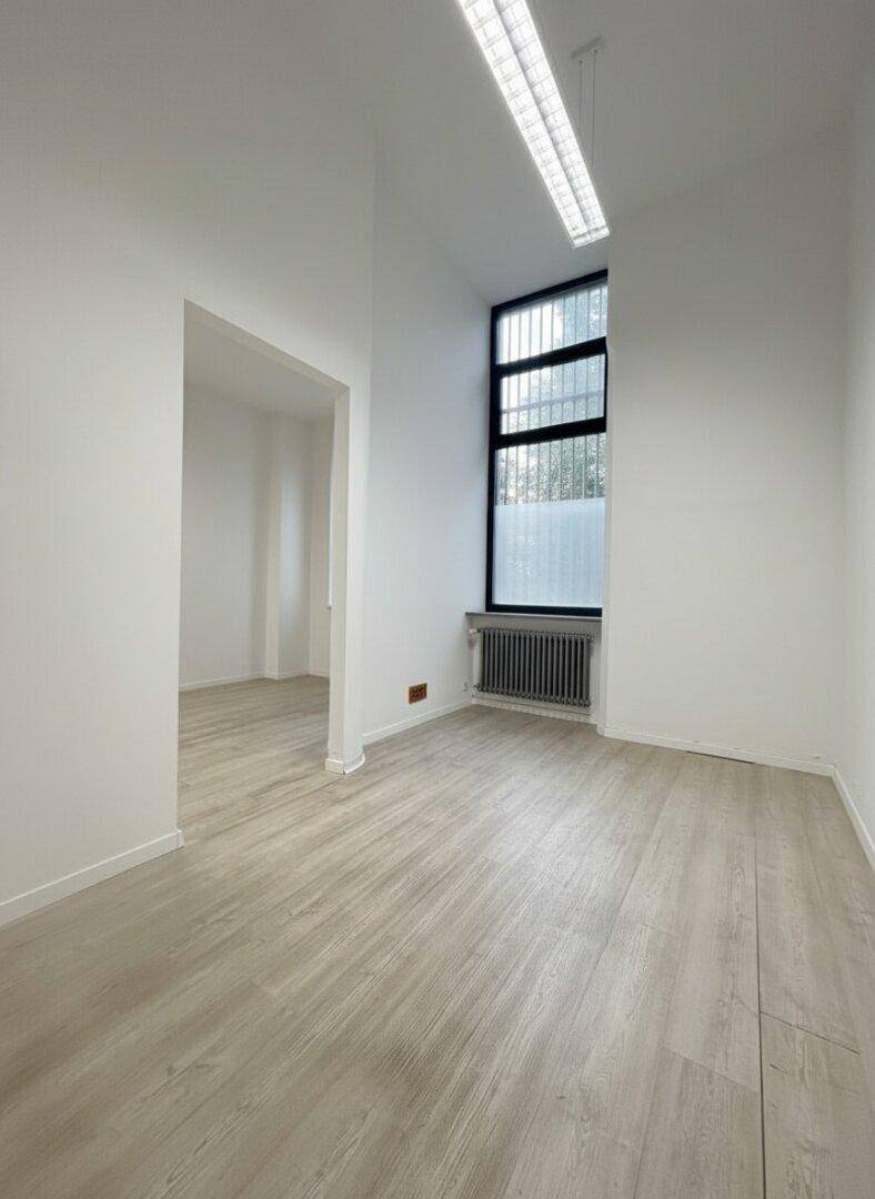 Leerer Büroraum mit hohen Decken, hellem Laminatboden und einem hohen Fenster, das Licht spendet.
