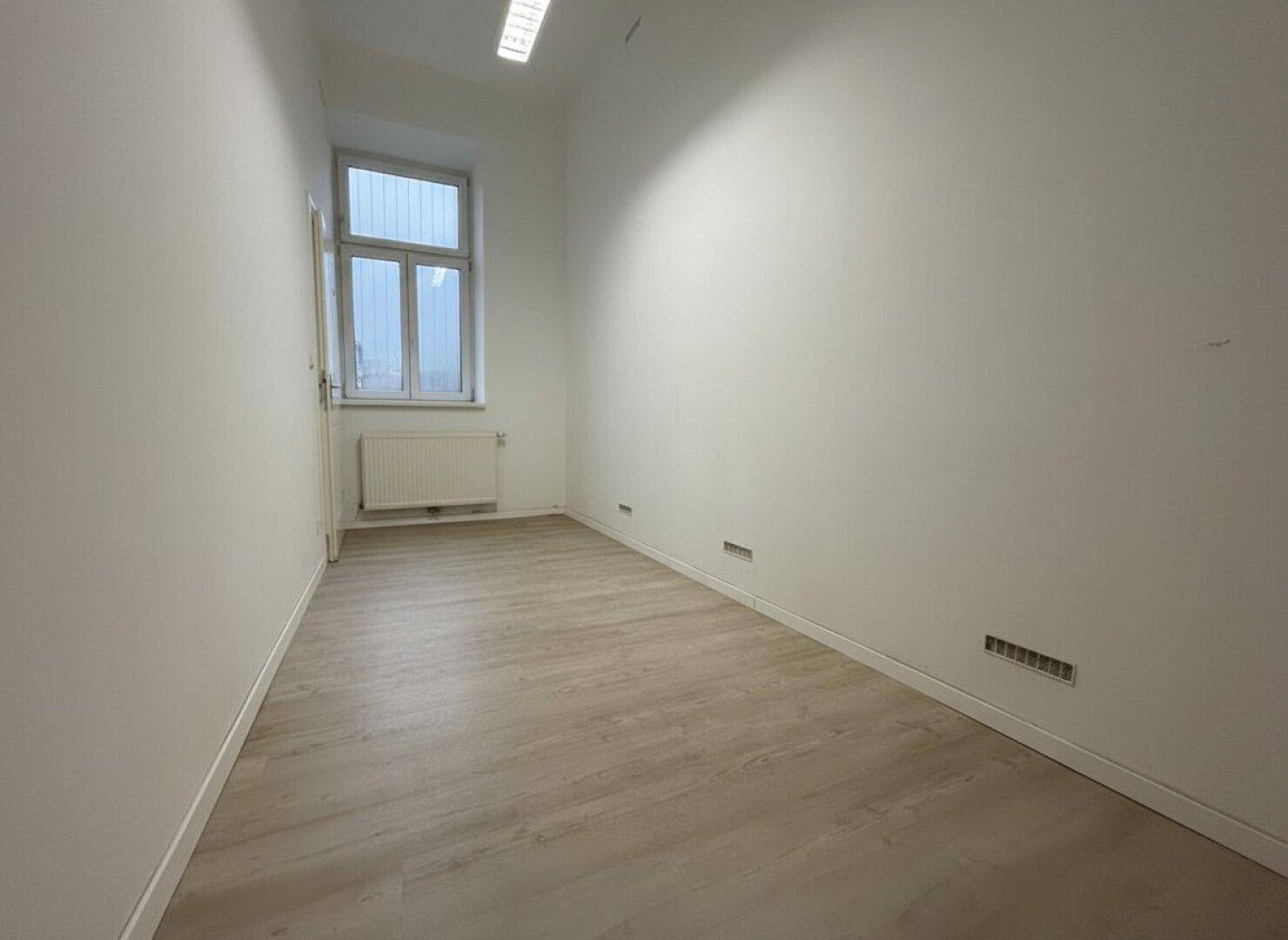 Schmaler Büroraum mit hellem Laminatboden, einem Fenster und weißen Wänden, ideal für Einzelbüro.