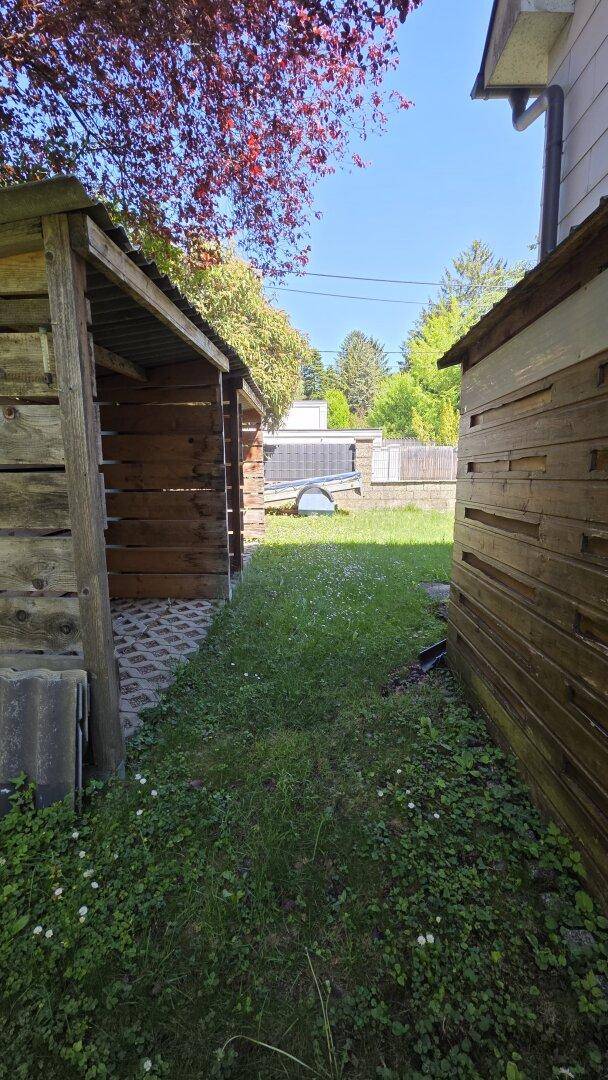 Schmaler Weg zwischen zwei Holzschuppen, der zu einer grünen Rasenfläche führt.