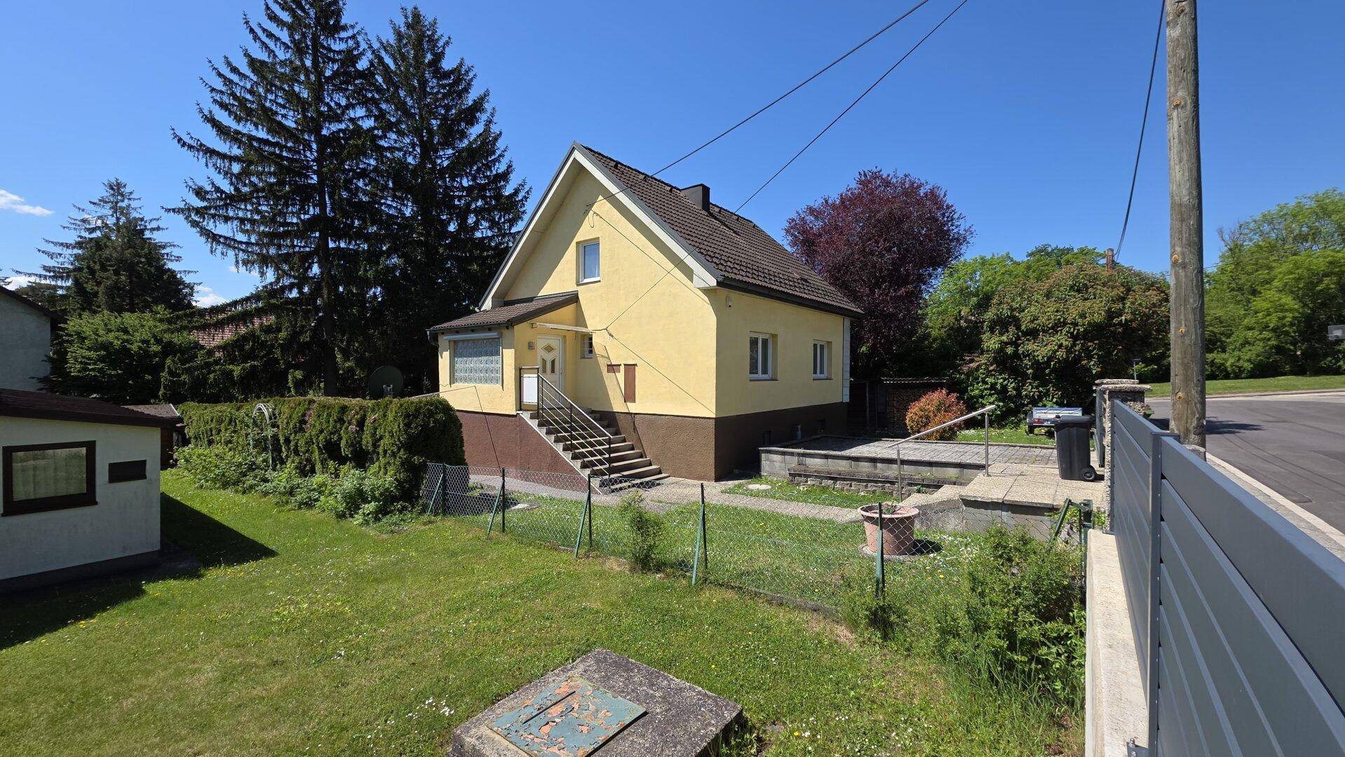 Gesamtansicht des gelben Hauses mit Vorgarten, Eingangsbereich und Zaun unter blauem Himmel.