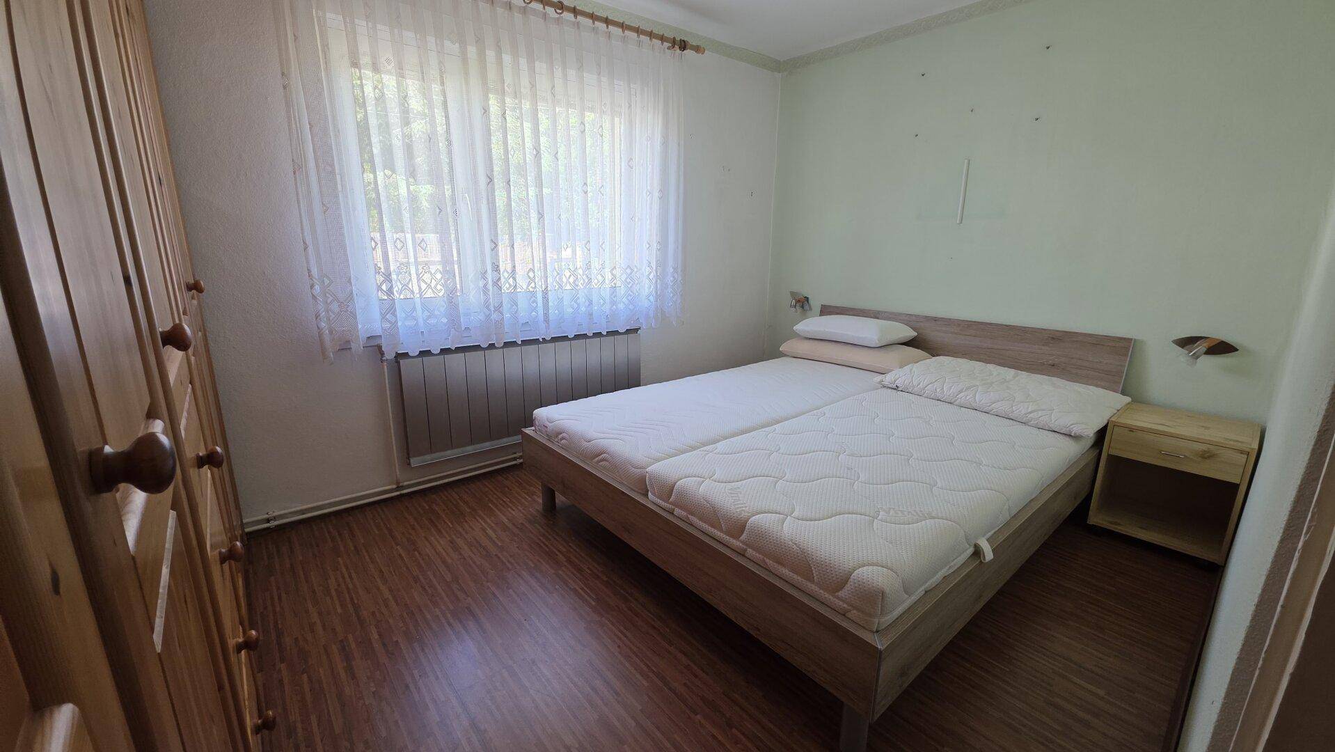 Helles Schlafzimmer mit Holzbett, Kleiderschrank und Fenster mit Vorhängen.