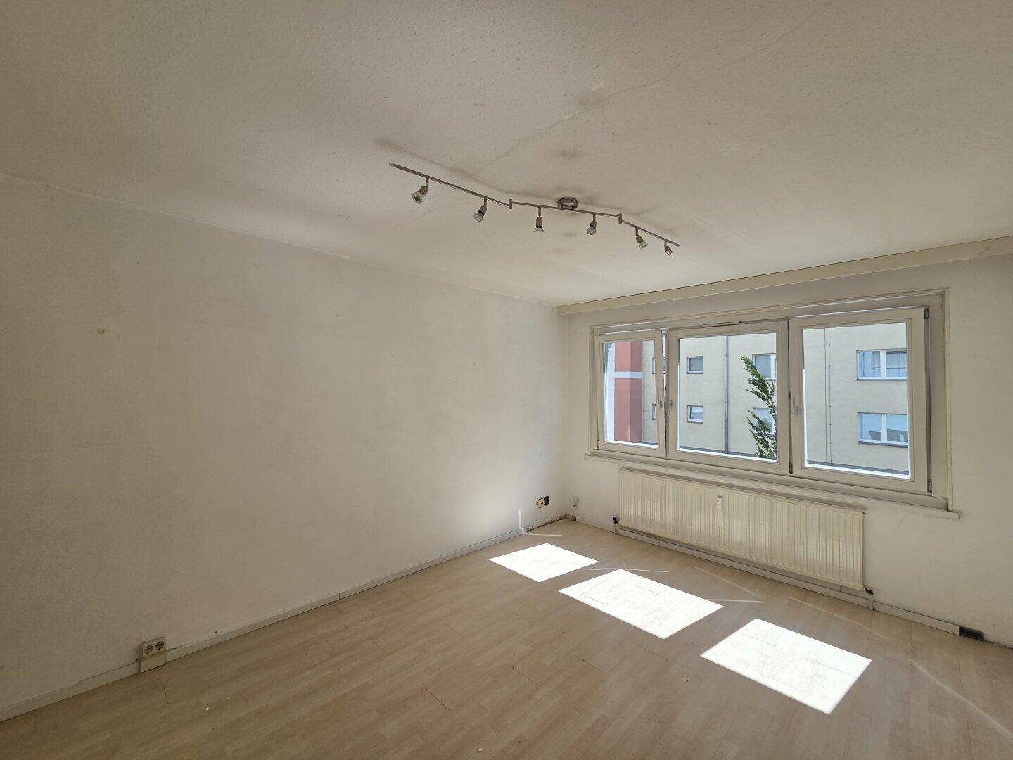 Leeres Wohnzimmer mit Laminatboden und großem Fenster, das viel Tageslicht hereinlässt.