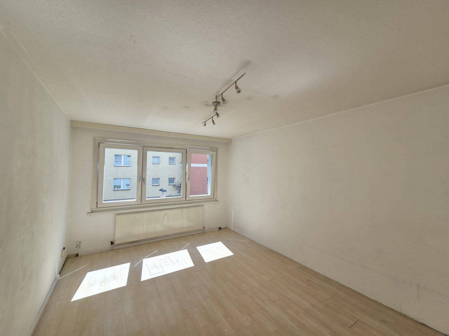 Leeres Schlafzimmer mit Laminatboden und großem Fenster, das viel Sonnenlicht hereinlässt.