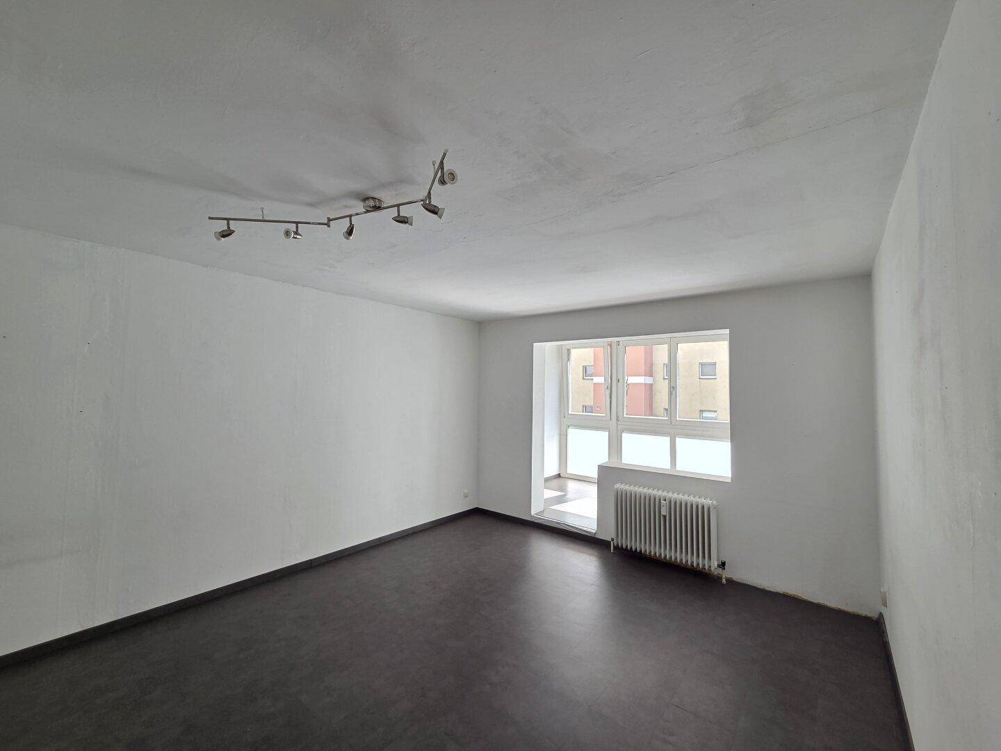 Leeres Wohnzimmer mit dunklem Boden und großem Fenster, das viel Tageslicht hereinlässt.