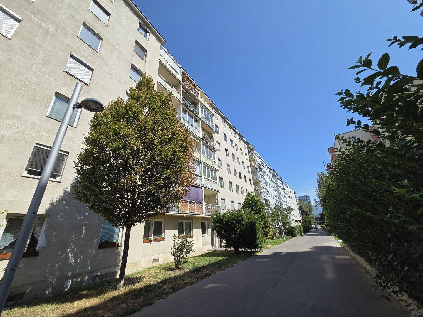 Mehrfamilienhausfassade mit Balkonen und Bäumen entlang eines Gehwegs an einem sonnigen Tag.