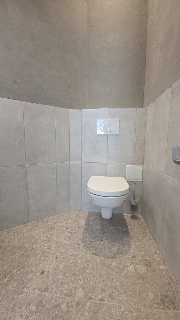 Modernes WC mit wandhängender Toilette und grauen Fliesen an Wänden und Boden.