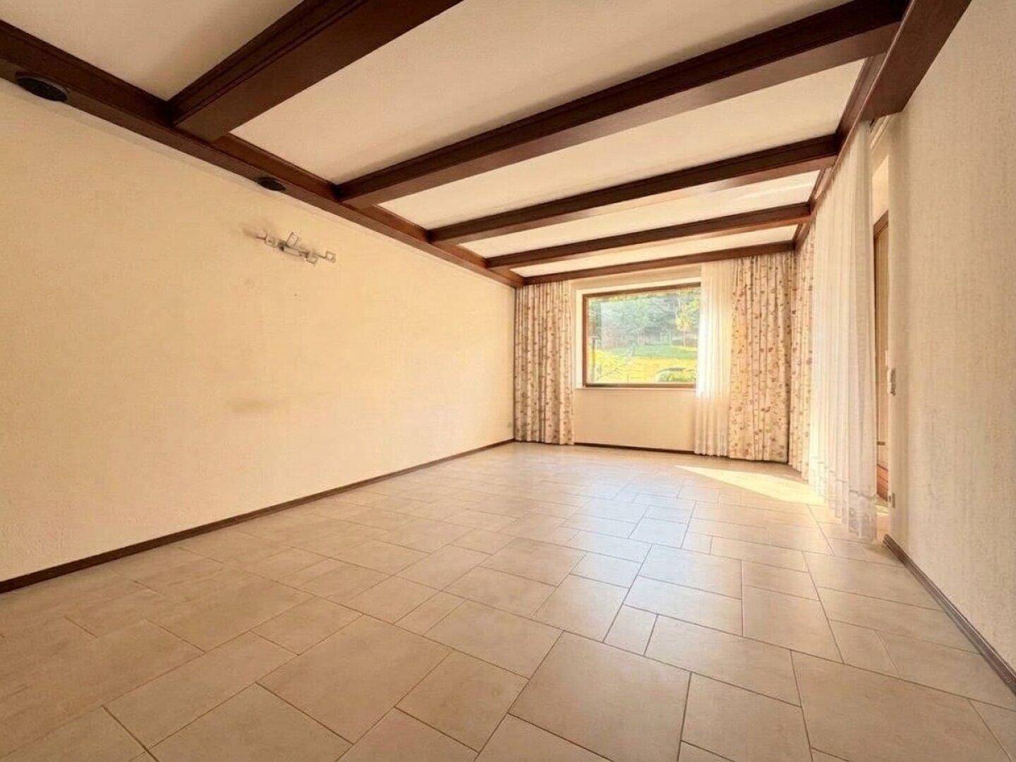 Geräumiges Wohnzimmer mit Fliesenboden, Holzbalkendecke und großem Fenster mit Gartenblick.