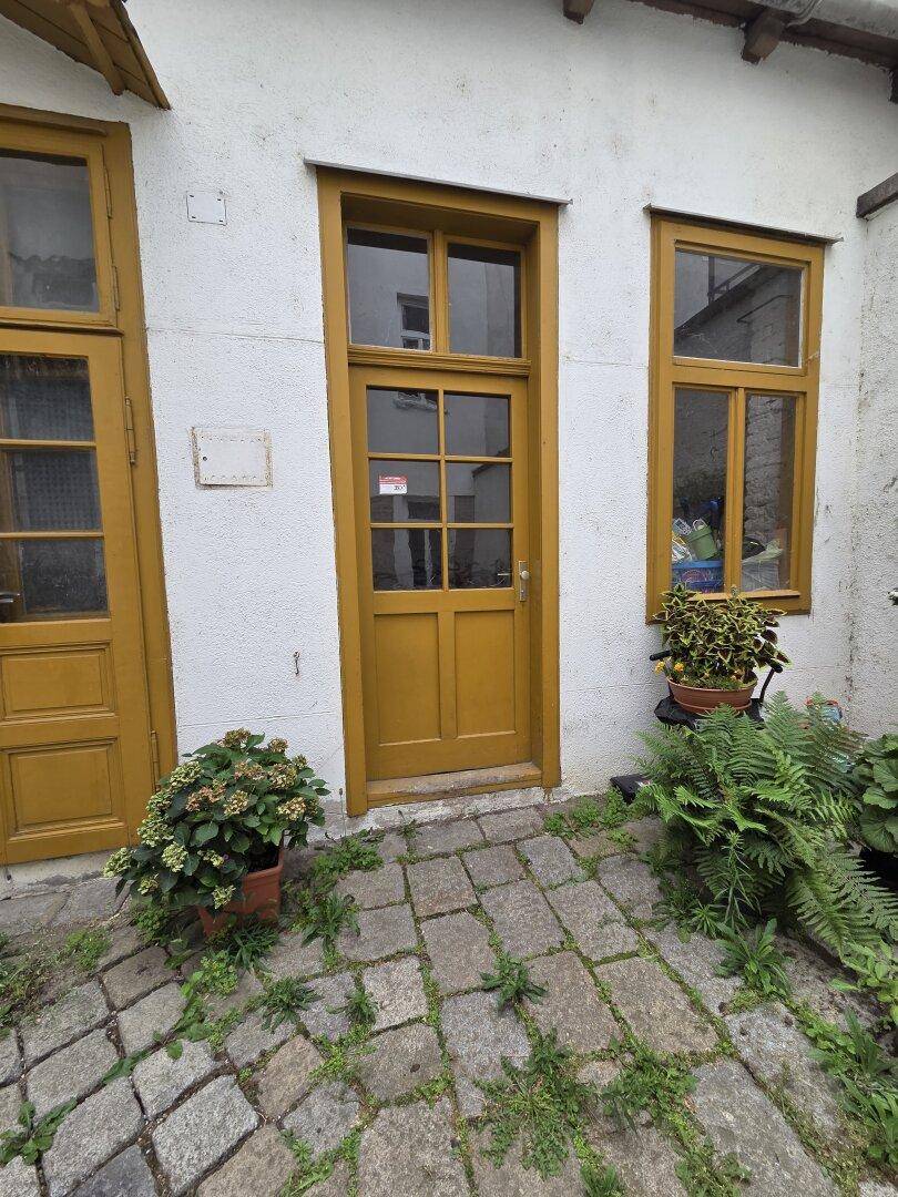 Eingangstür mit gelbem Rahmen und Fenstern, umgeben von Pflanzen im gepflasterten Innenhof.