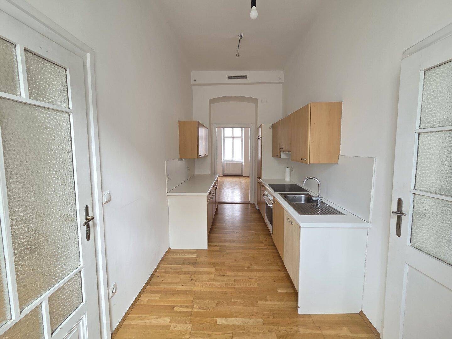 Schmale, funktionale Küche mit hellen Holzschränken, weißen Arbeitsplatten und Parkettboden.