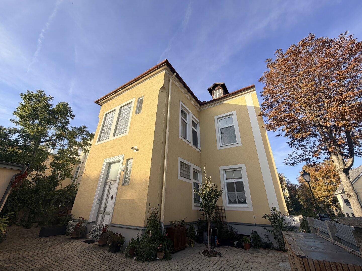 Seitenansicht eines gelben Gebäudes mit vielen Fenstern und gepflastertem Vorplatz.
