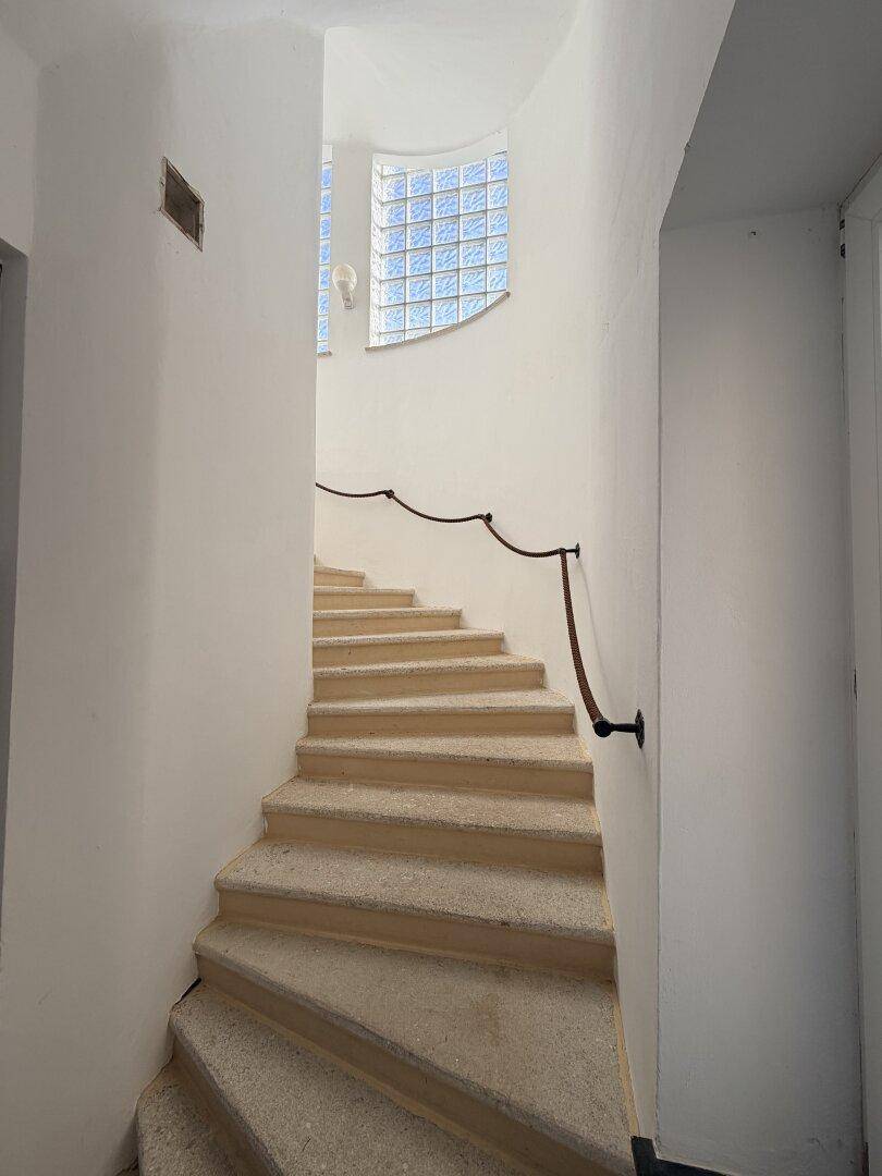 Geschwungene Steintreppe mit Geländer in einem hellen Treppenhaus mit Glasbausteinen.