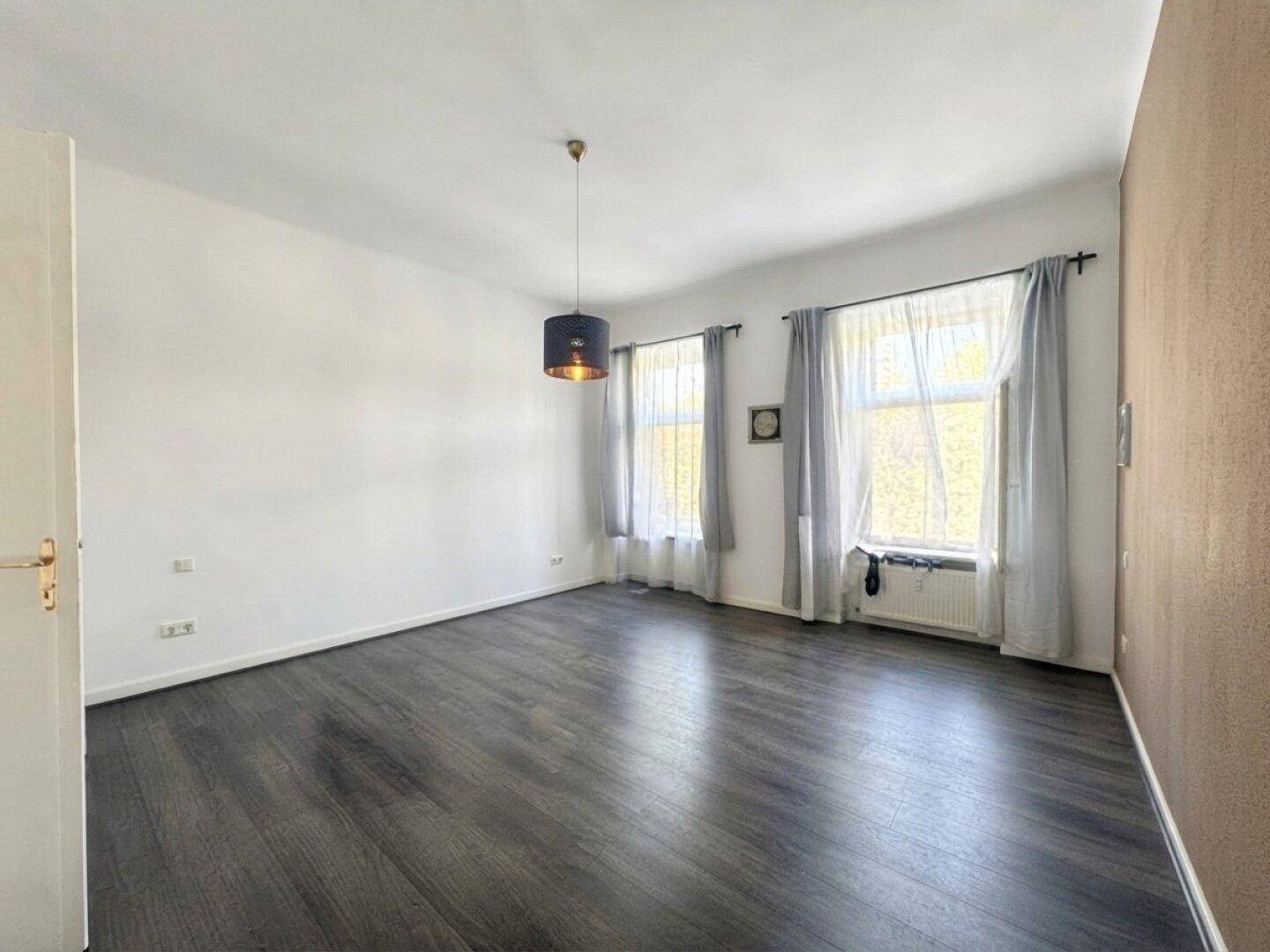 Geräumiges Wohnzimmer mit dunklem Laminatboden und zwei Fenstern, bereit zur Einrichtung.