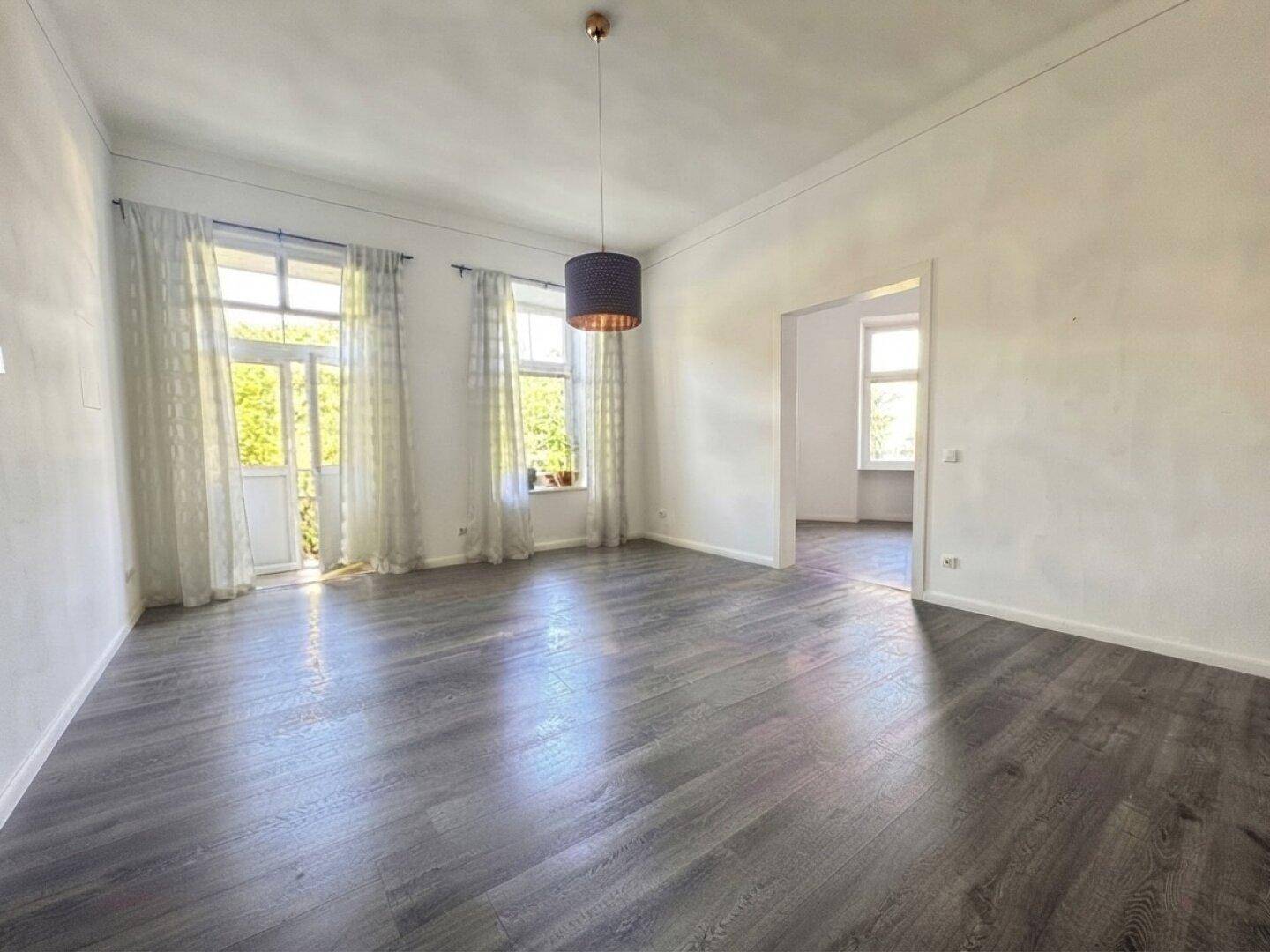 Helles, leeres Wohnzimmer mit dunklem Laminatboden und Zugang zu einem Balkon.