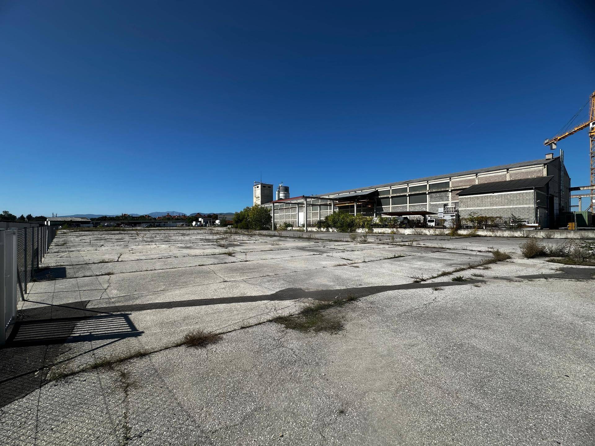 Weitläufige, gepflasterte Außenfläche mit einem Industriegebäude und einem Kran im Hintergrund unter blauem Himmel.