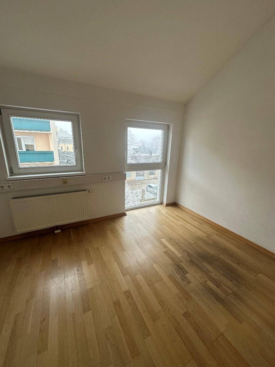 Helles Zimmer mit Holzboden, weißen Wänden und Blick auf die umliegenden Gebäude und verschneite Bäume.