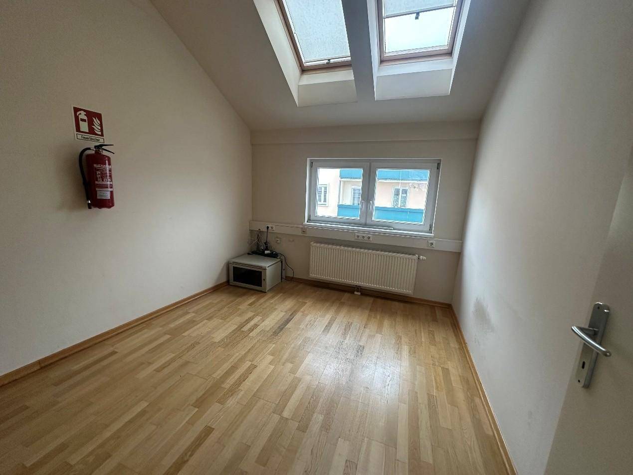 Helles Zimmer mit Holzboden, Dachfenstern und einem Heizkörper, ausgestattet mit einem Feuerlöscher.