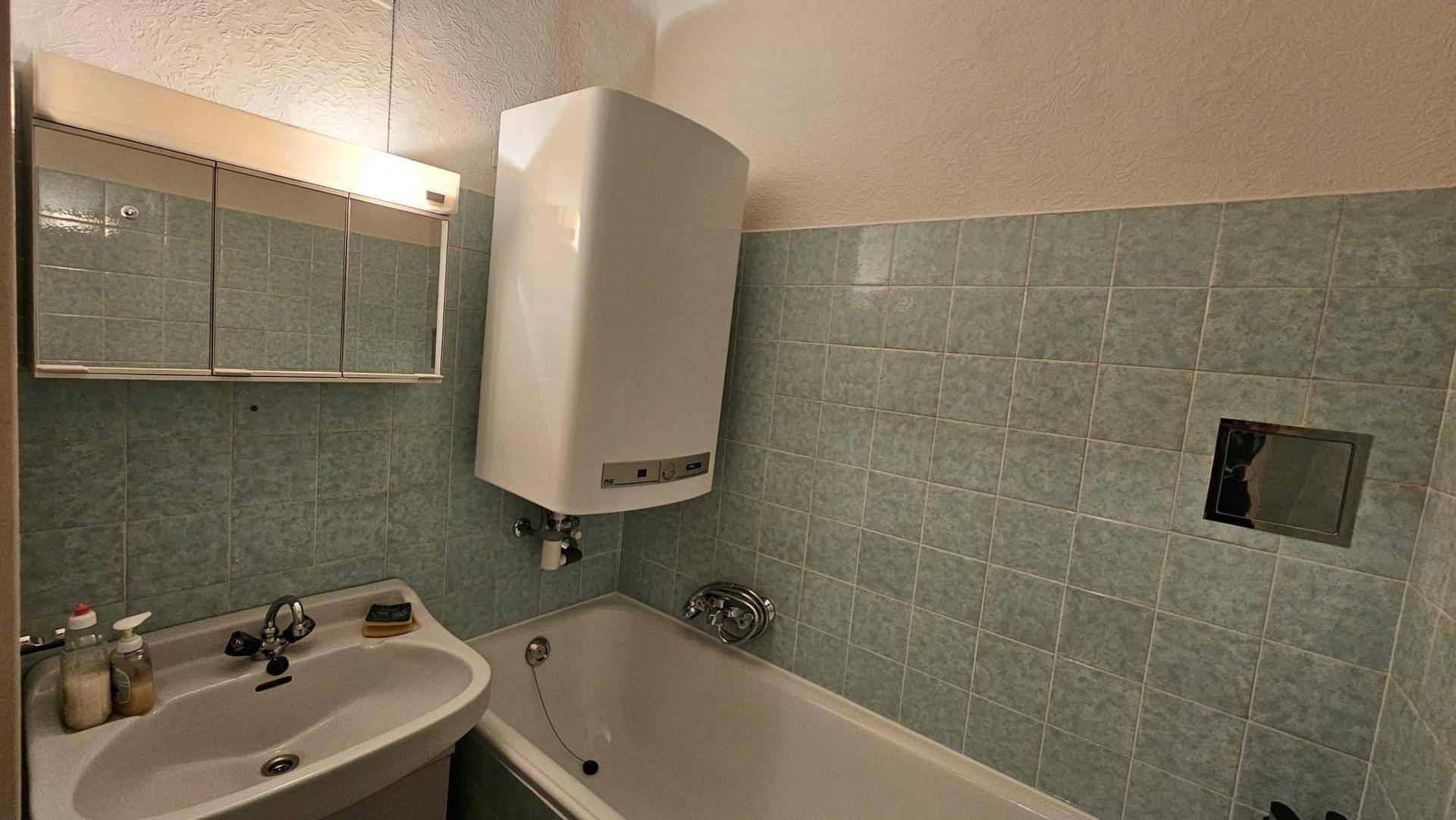 Badezimmer mit Badewanne, Waschbecken, Spiegelschrank und hellgrünen Fliesen im 80er-Jahre-Stil.