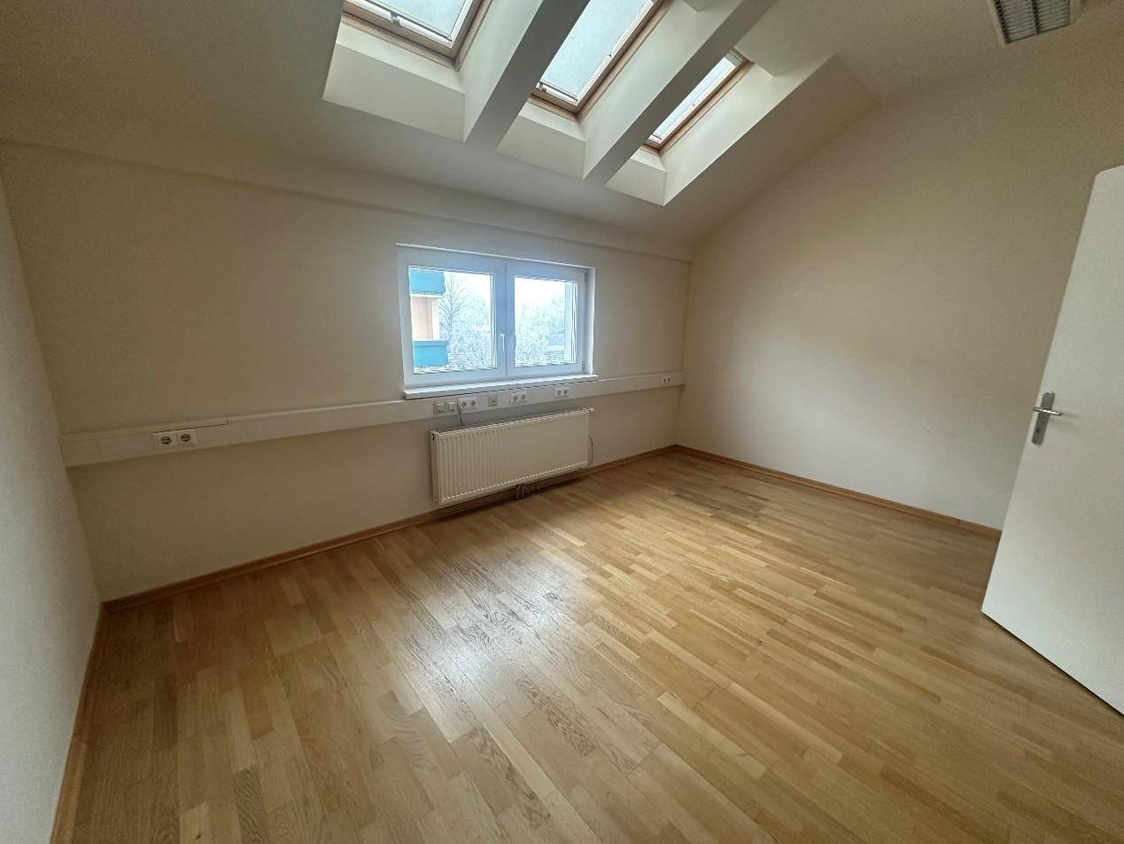 Helles Zimmer mit Holzboden, Dachfenstern und einem Heizkörper, das viel Tageslicht durch die Fenster erhält.