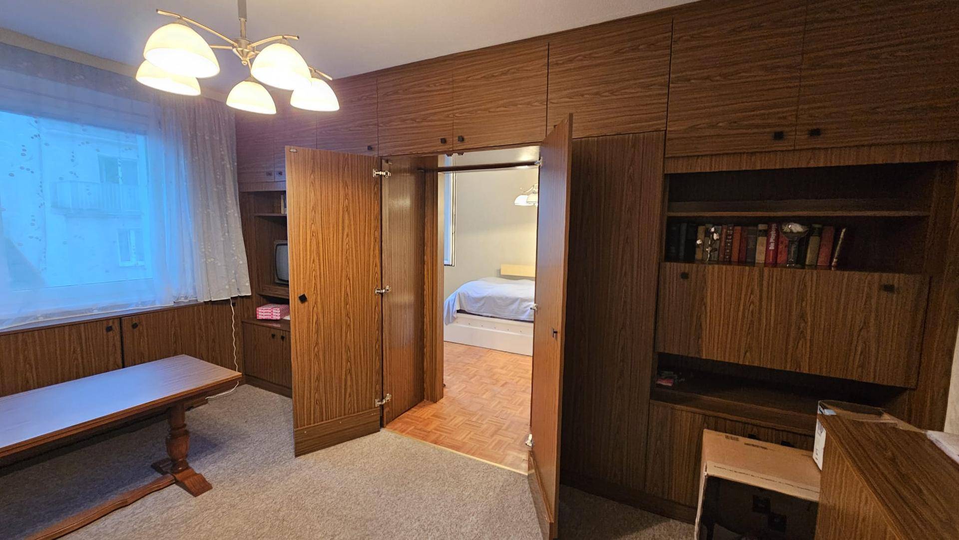 Wohnzimmer mit Holzvertäfelung und offener Tür zu einem angrenzenden Schlafzimmer mit Parkettboden.