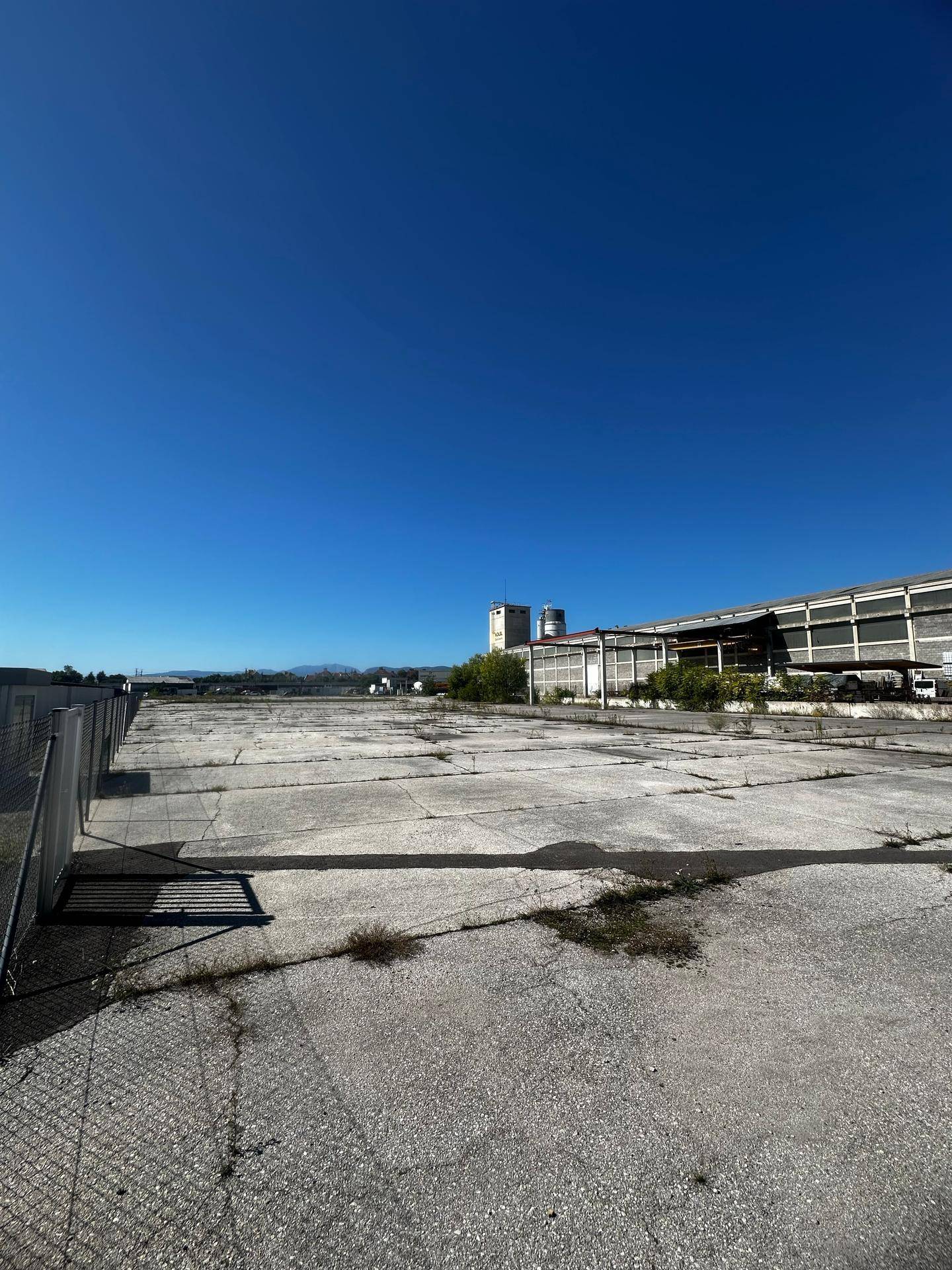 Ausgedehnte, sonnige Außenfläche mit einem Industriegebäude und einem Zaun im Vordergrund unter klarem Himmel.