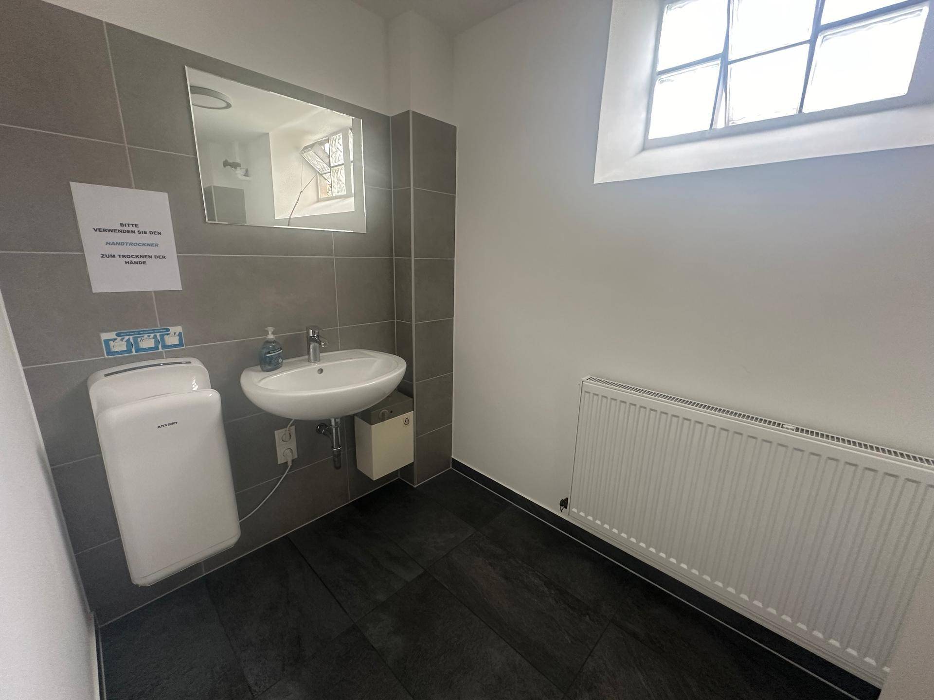 Modernes Badezimmer mit grauen Fliesen, Waschbecken und Händetrockner, sauber und funktional.