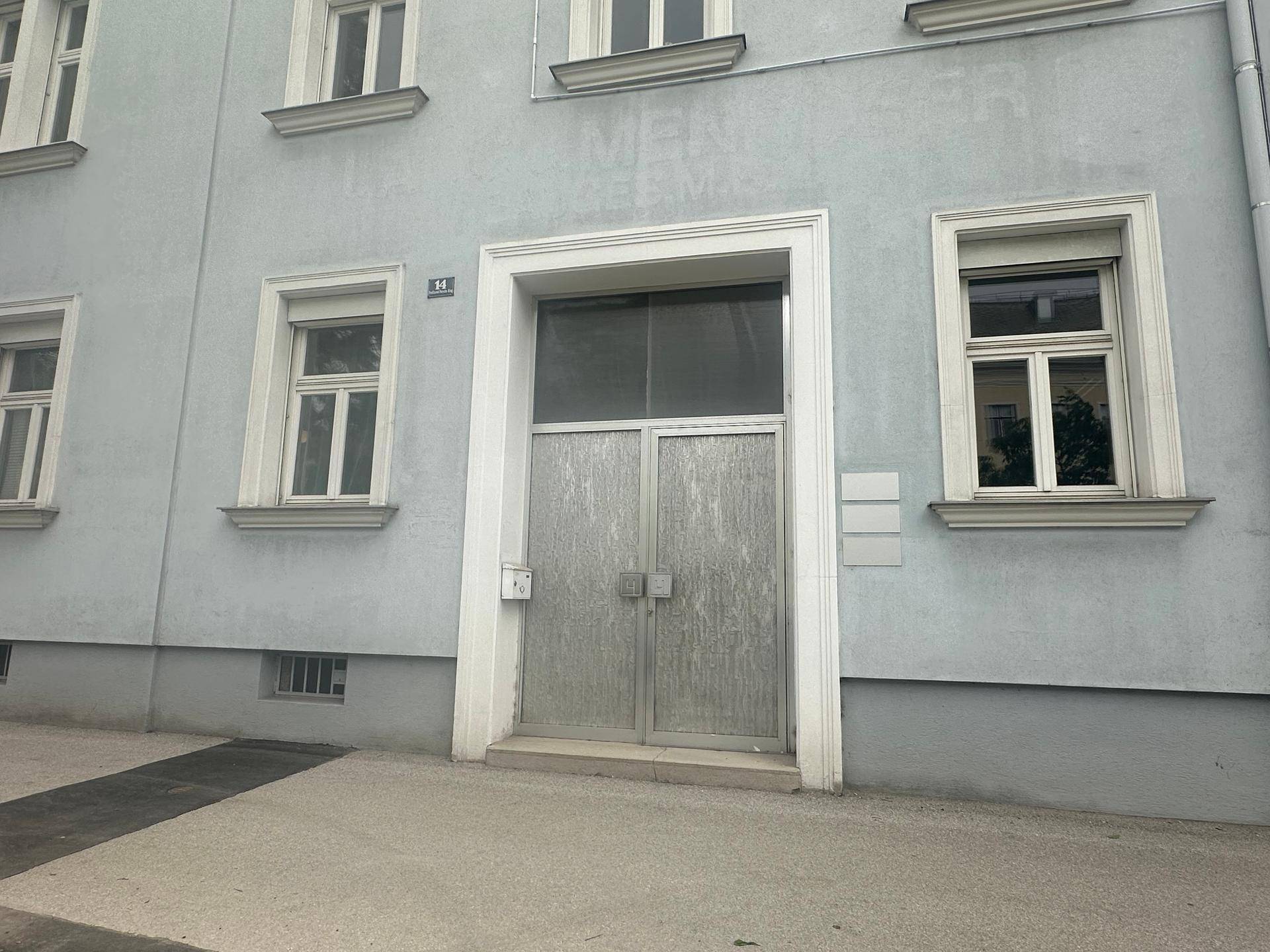 Fassade eines Gebäudes mit großem Eingangstor und mehreren Fenstern, in hellem Blaugrau gestrichen.