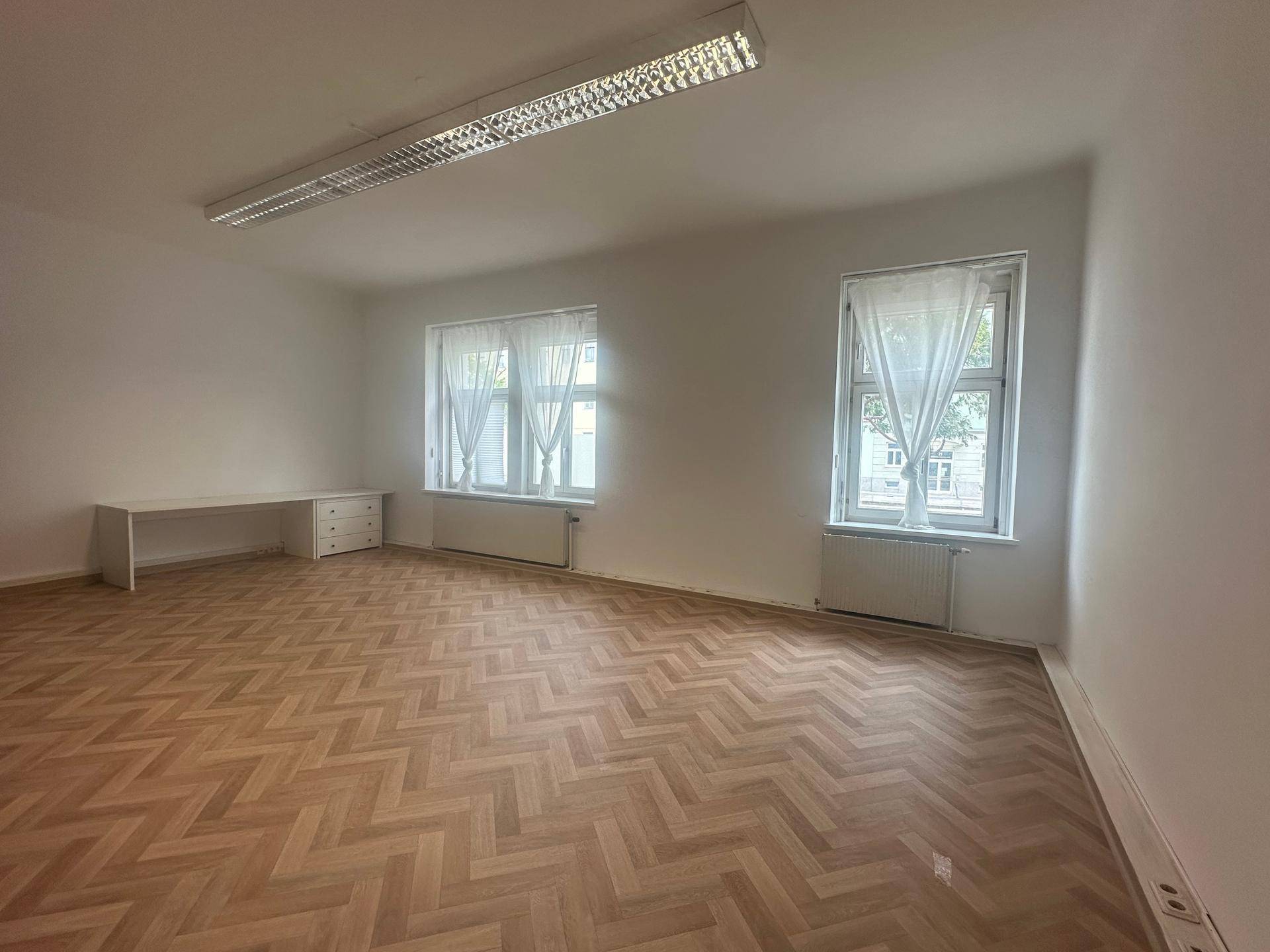 Freundlicher Büroraum mit Fischgrätparkett, Schreibtisch und großen Fenstern für eine angenehme Arbeitsatmosphäre.