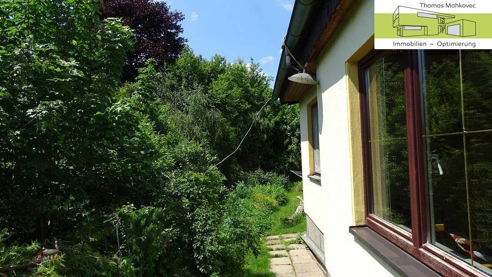 Seitenansicht des Hauses, umgeben von dichtem Grün und Bäumen.