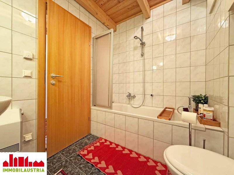 Badezimmer mit Badewanne und Duschvorrichtung, weißen Fliesen und Holzbalkendecke.