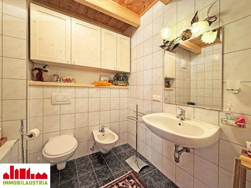 Badezimmer mit weißen Fliesen, Waschbecken, Toilette und Bidet unter der Holzbalkendecke.