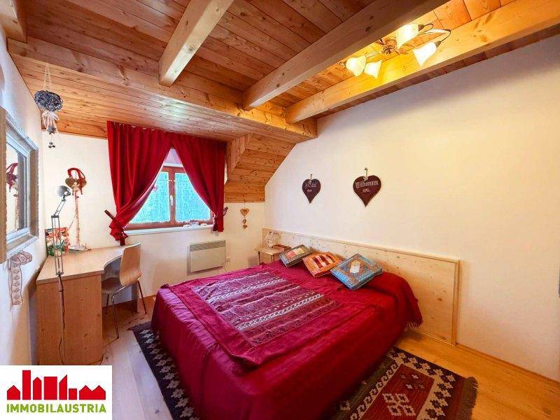 Schlafzimmer mit Holzbett, Schreibtisch und Dachschräge, dekoriert mit roten Vorhängen.