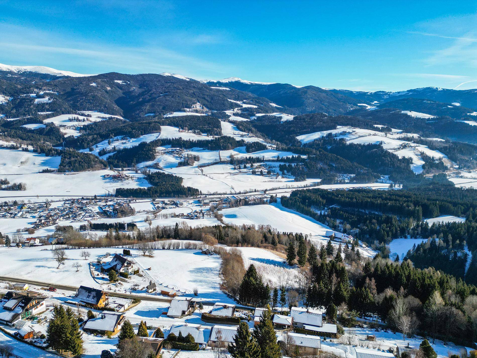 Weitläufige Luftaufnahme der verschneiten Berglandschaft mit verstreuten Häusern und Wäldern.