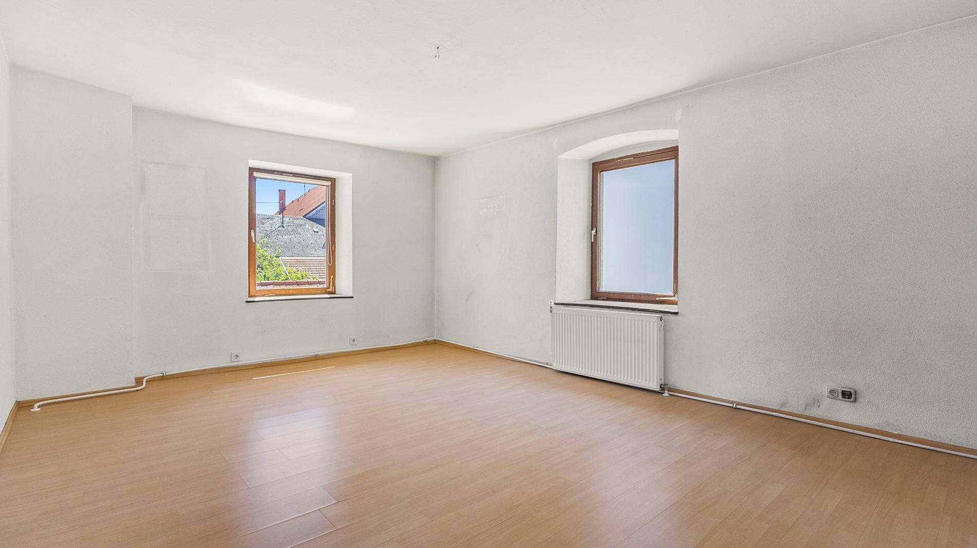 Leeres Schlafzimmer mit Holzboden, weiß gestrichenen Wänden und Heizkörper unter dem Fenster.
