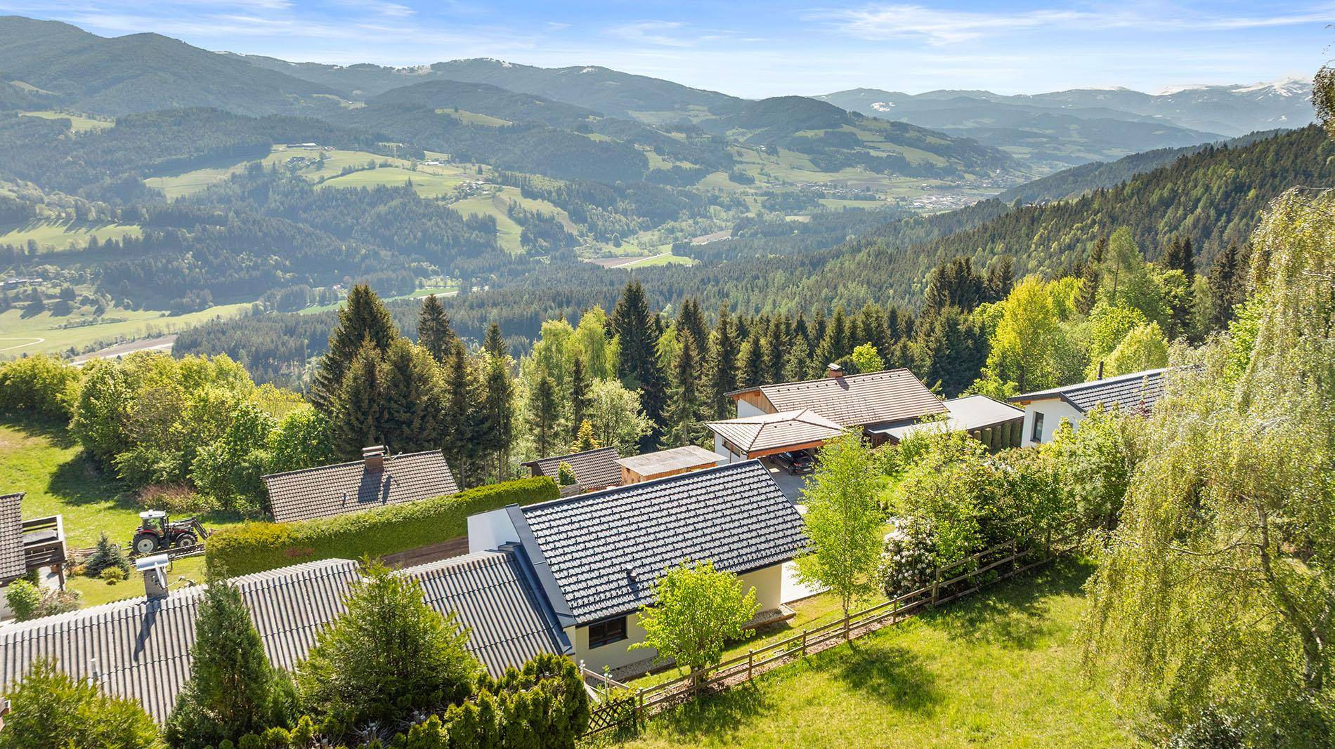 Vogelperspektive des Hauses mit dunklem Dach, umgeben von grünen Bäumen und Bergen.