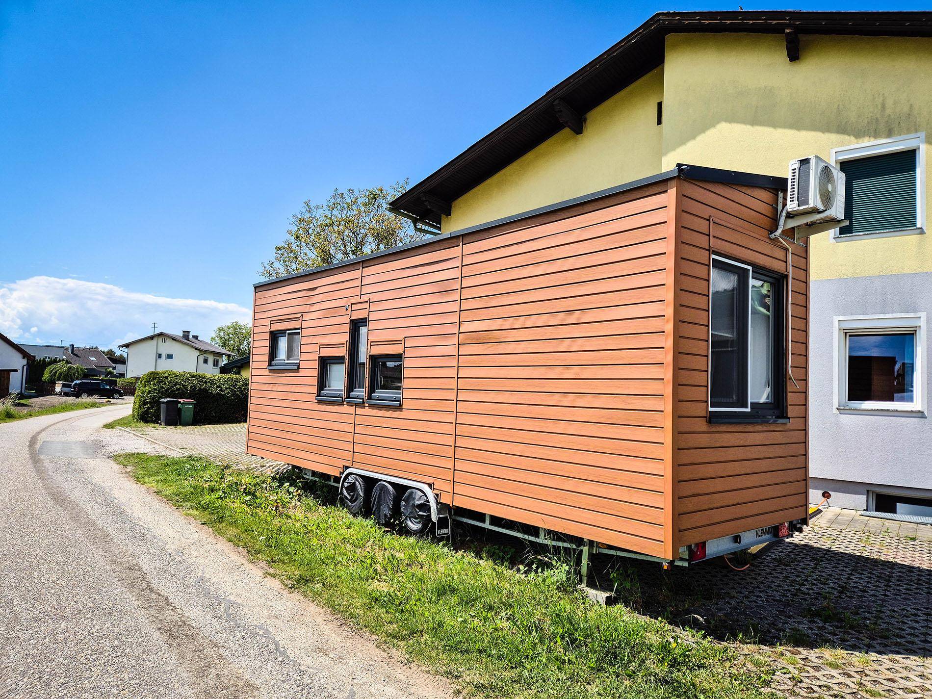 Seitliche Ansicht des Tiny Houses mit Holzfassade, geparkt neben einem Wohnhaus.