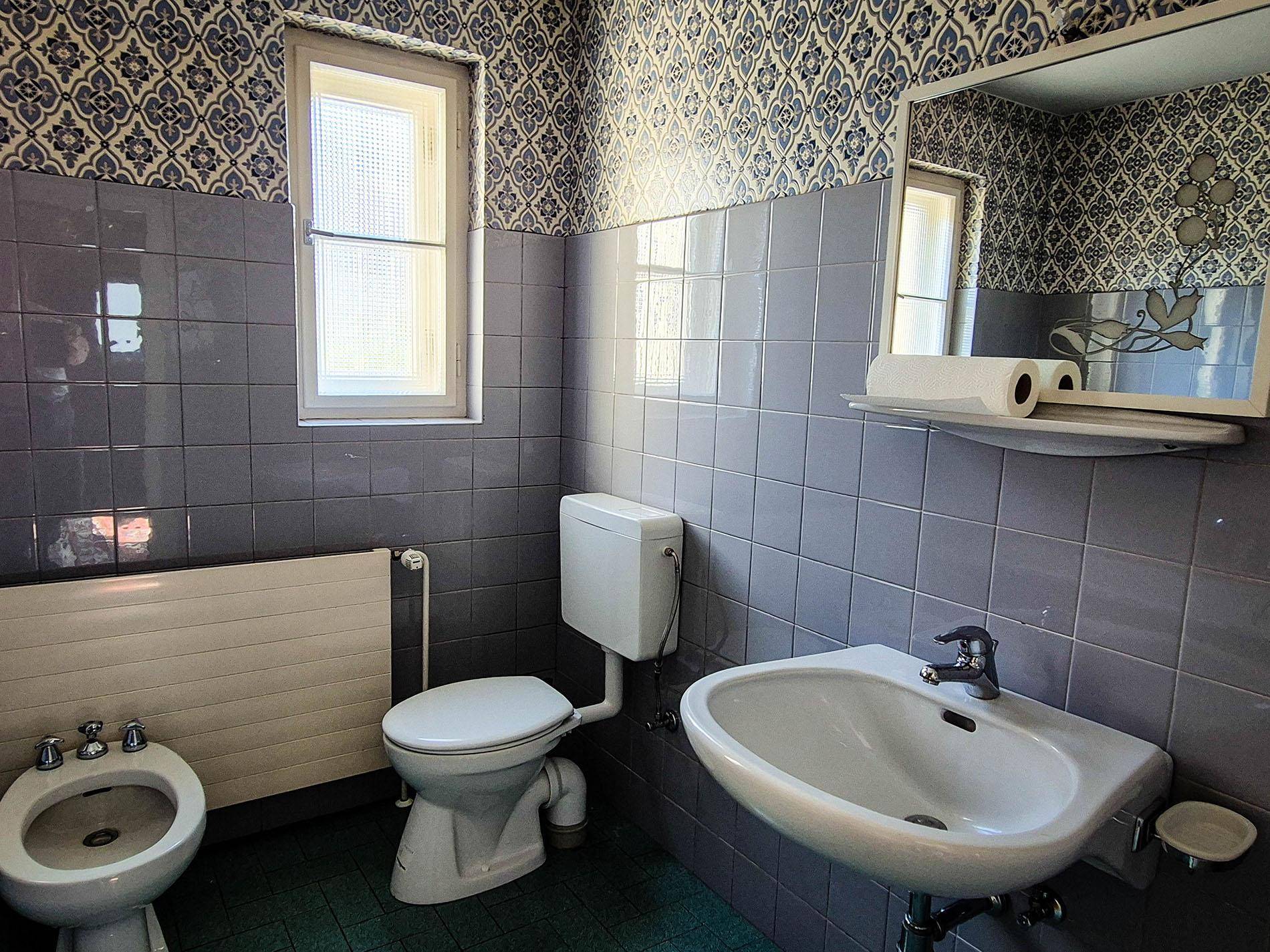 Badezimmer mit blaugrauen Fliesen, WC, Waschbecken, Bidet und Fenster.