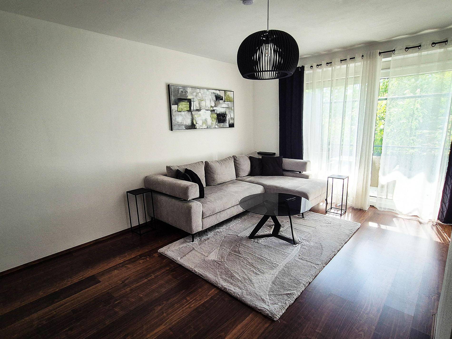 Helles Wohnzimmer mit großem Ecksofa, dunklem Holzboden und Fenstern mit Vorhängen.