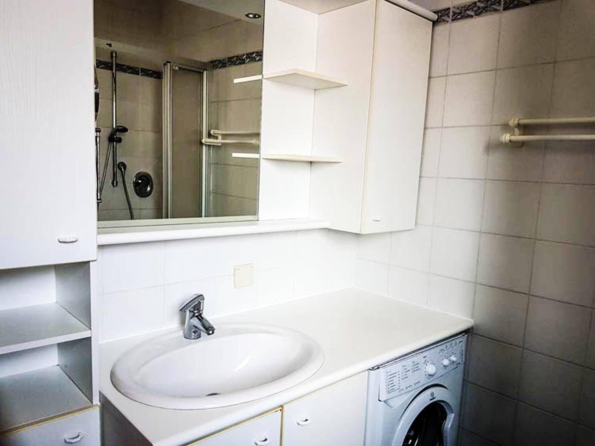 Badezimmer mit weißen Fliesen, Waschbecken, Waschmaschine und einer Duschkabine.