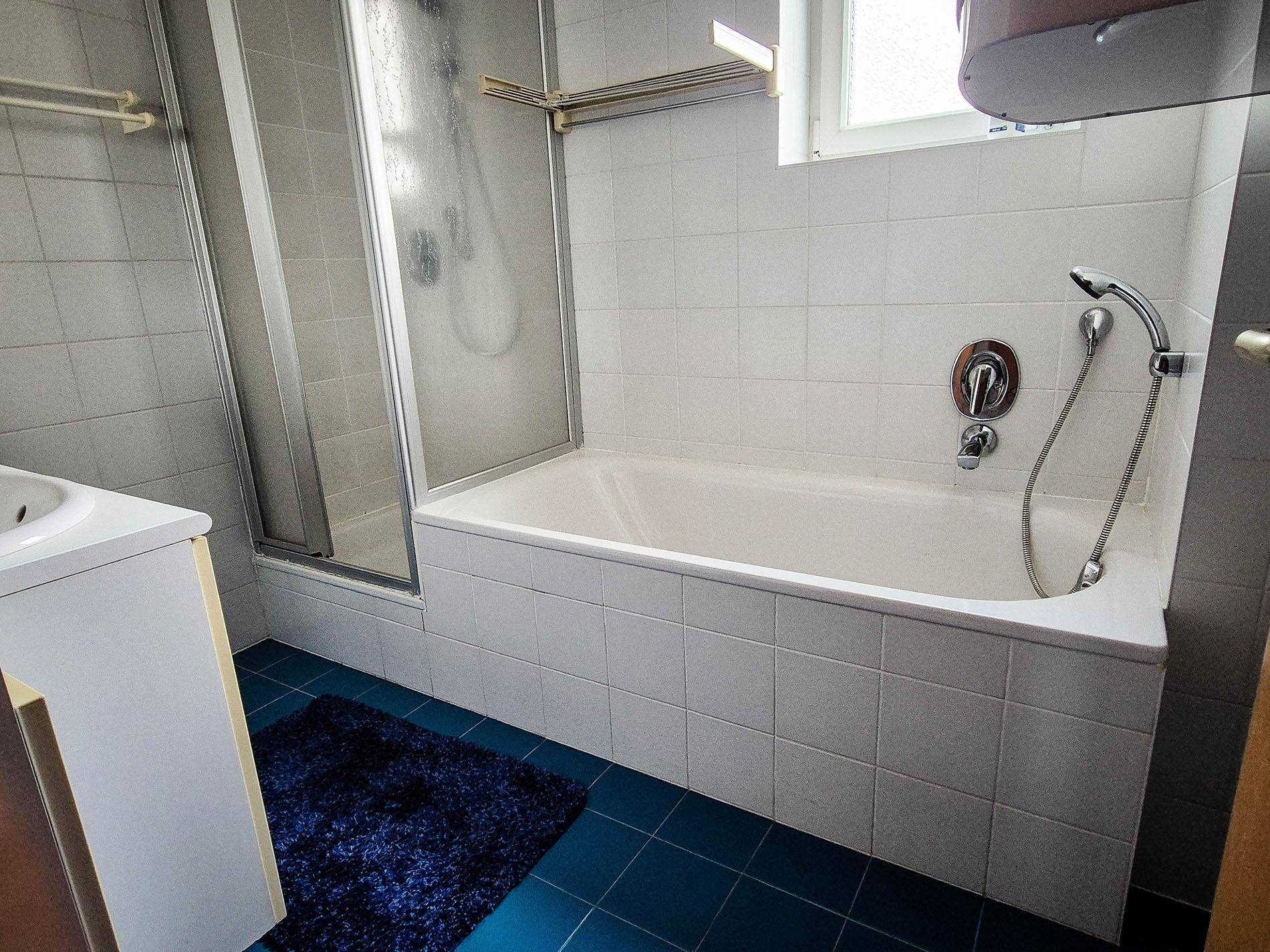 Badezimmer mit Badewanne, Duschbereich und blauen Bodenfliesen sowie weißen Wandfliesen.
