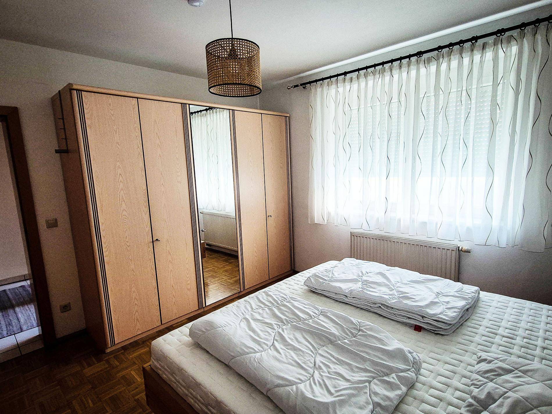 Schlafzimmer mit Doppelbett, großem Holzschrank mit Spiegeltüren und Fenster.