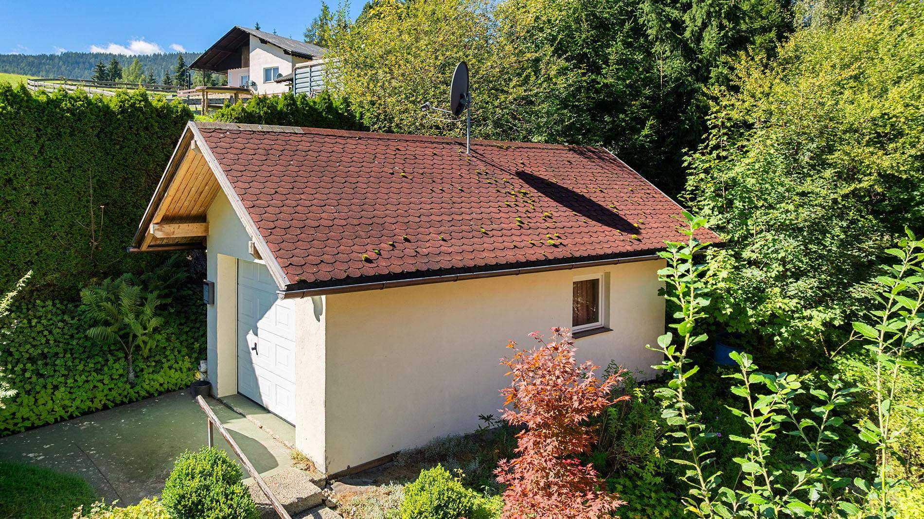 Ansicht einer hellen Garage mit rotem Satteldach, umgeben von grüner Hecke und Bäumen.