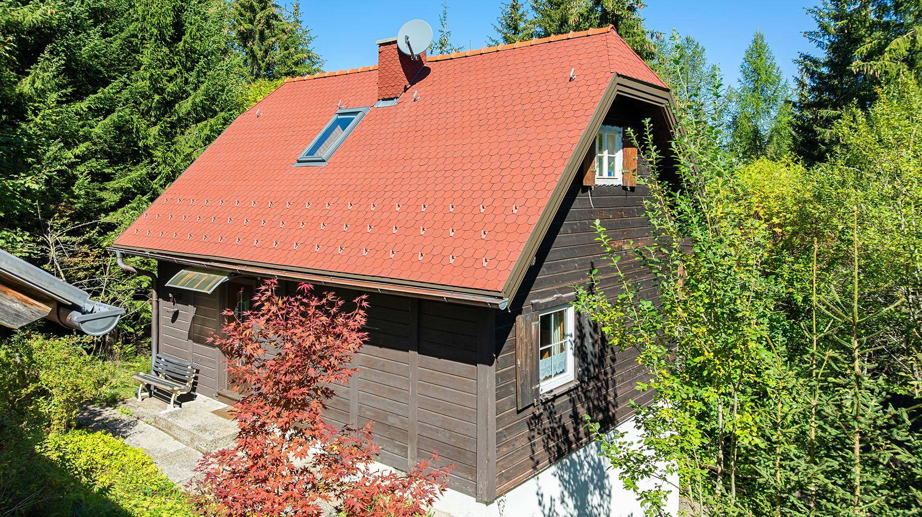 Nahaufnahme eines Holzhauses mit rotem Satteldach und Dachfenster, umgeben von Bäumen.