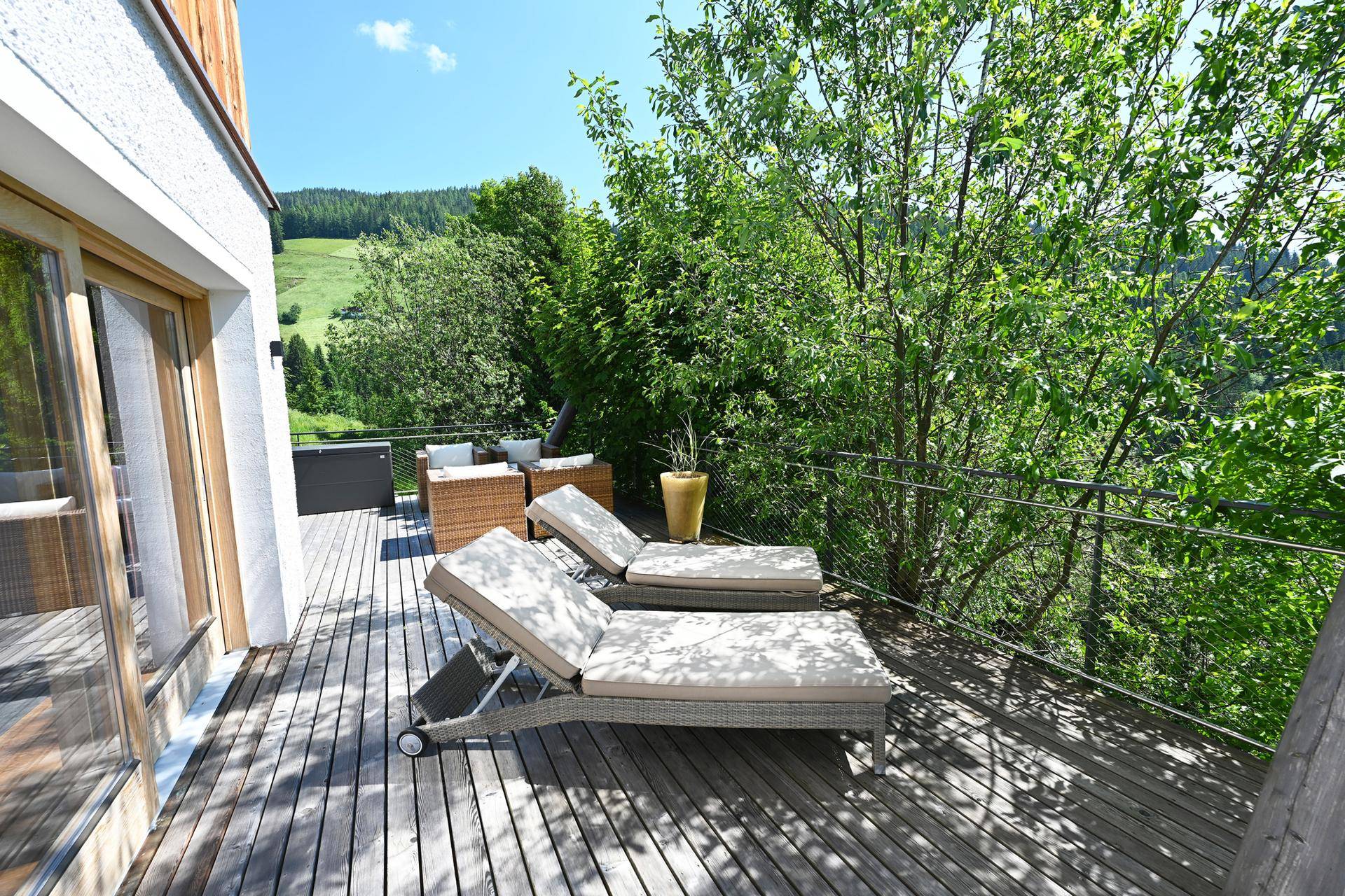 Sonnige Holzterrasse mit Liegestühlen, umgeben von Bäumen und mit Blick auf die grüne Landschaft.