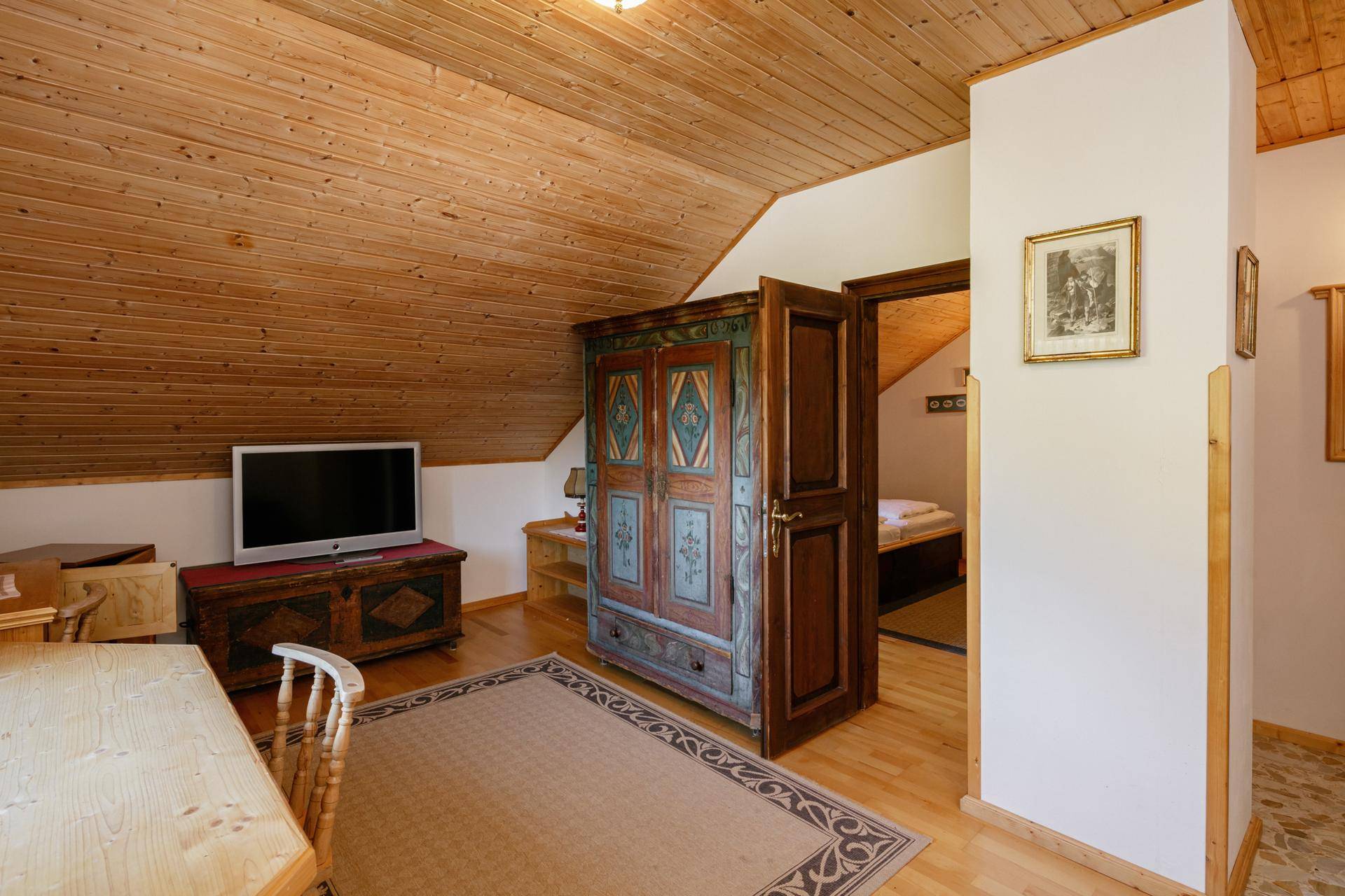 Schlafzimmer mit Holzboden, schrägen Decken und einem antiken Schrank, der zu einem weiteren Raum führt.